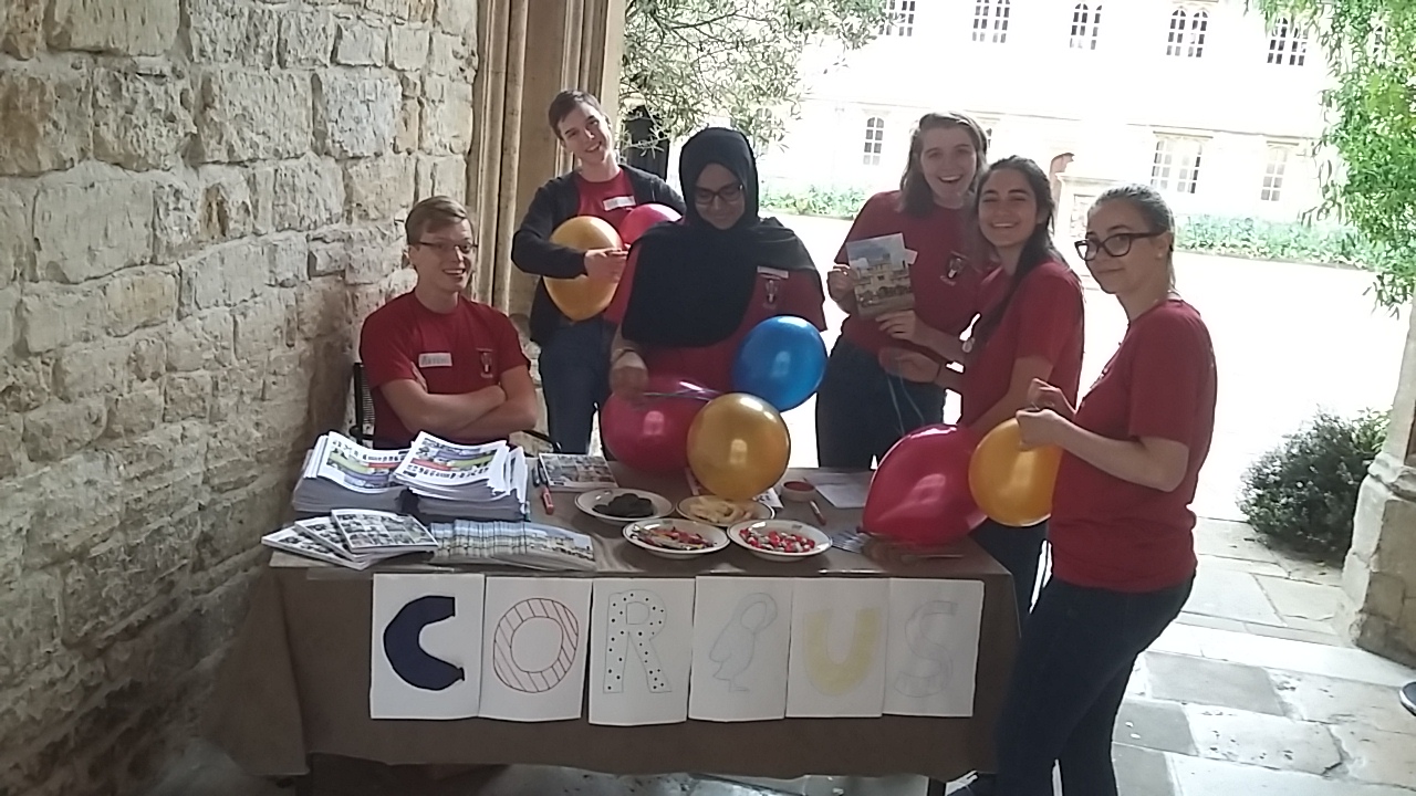 Corpus Christi College's front desk on Open Day. There are six students holding balloons and standing next to a table which has 'Corpus' written on the front.