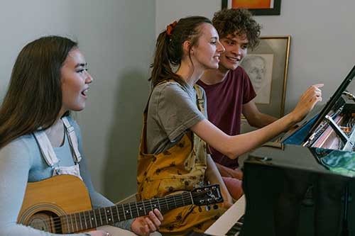 Three students sitting at a piano, one holding a guitar