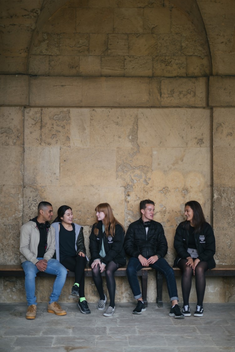 Five students sitting on a bench in front a stone wall in Worcester College