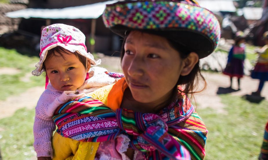A young Peruvian woman wearing a colourful hat and shawl holds a baby. She is standing outside. Village buildings are visible behind her.