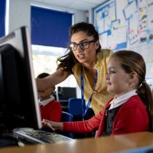 A close-up front view of a teacher and her young pupils who are taking part in a computing class. The teacher is offering one of the girls in her class some advice as she gets to grip with the computer.