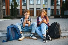 Two children looking at mobile phones sitting in a public space. Credit: Adobe Stock