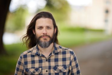 A portrait photograph of Aleks Kissinger, a middle-aged white man with long brown hair and a beard, wearing a checked shirt.