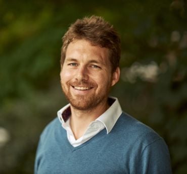 A white man with short brown hair and a beard, wearing a white shirt and blue jumper. He stands in front of an out-of-focus hedge backdrop.