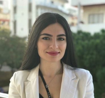 Dr Amani Maalouf. She wears a smart jacket. There are buildings and trees in the background behind her.