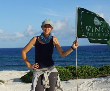 A white woman stands by an ocean holding a flag which has the words ‘Wings Worldquest’ 