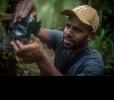 A young Indonesian map wearing a T shirt and baseball hat adjusts a camera in the middle of the jungle.