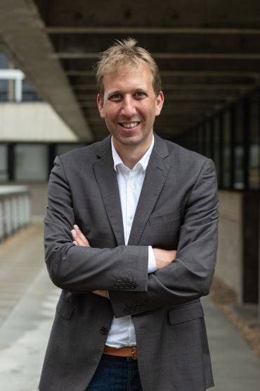Chris Lintott, a white man with short brown hair wearing a grey jacket and white shirt. He stands with his arms folded outside a university department building. 