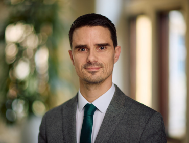 A white man with short dark hair wearing a grey suit and emerald green tie.