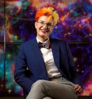 Portrait of Dr H&eacute;lo&iuml;se Stevance, a young lady with curly orange and red hair, wearing a dark blue jacket, glasses and a colourful bow tie. She sits in front of a colourful outer-space backdrop.