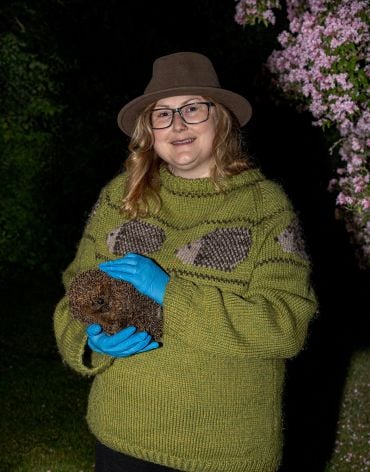 A lady with long blonde hair wearing a green jumper and a brown hat holds a hedgehog in an outdoor area at night.