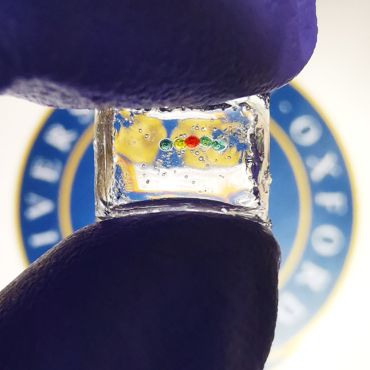 A chain of five coloured droplets is suspended in a cube of clear jelly-like material, held between a thumb and forefinger (wearing a latex glove). Through the cube, the University of Oxford’s logo can be seen, demonstrating the transparency of the cube.