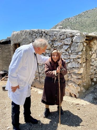 A man wearing a white coat stands with an elderly lady wearing a hooded coat and holding onto a staff, outside a stone dwelling.