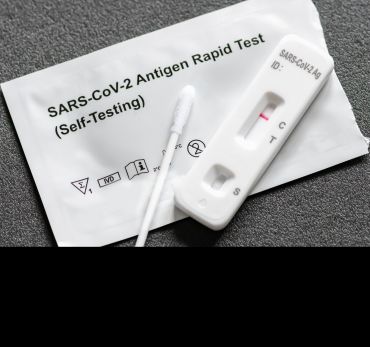 A Covid-19 antigen test kit. A swab lies on top of a test strip (showing a negative result). The opened packet for the test is in the background.