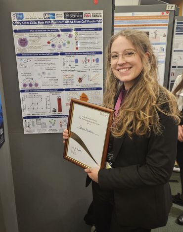 A smiling young lady with long dark blonde hair and glasses holds a certificate. She stands in front of a large poster which summarises her scientific research.