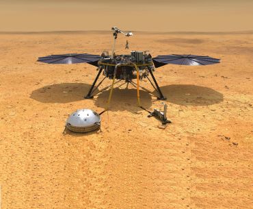 A spacecraft with a tripod structure on the surface of Mars, seen as an arid, desert-like landscape. The spacecraft has an arm-like probe that extends upwards, and two solar panels on either side, which stick out like wings.