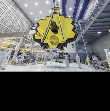 The James Webb Space Telescope’s gold mirror is made up of hexagonal sections joined together. It is suspended in a large warehouse, with several people in the foreground, demonstrating the large size of the mirror. 