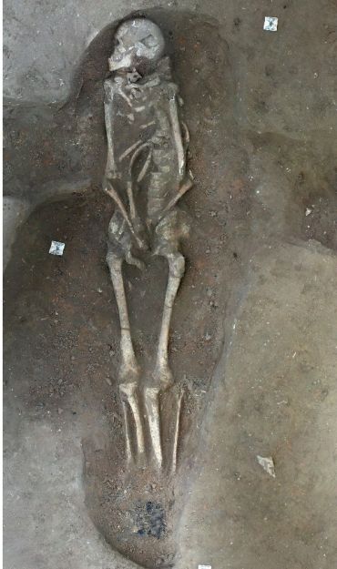 An excavated human skeleton lies at the bottom of a deep oval-shaped pit.