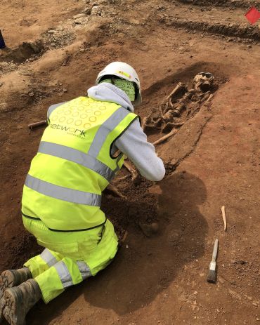 A person wearing a helmet, a fluorescent jacket and fluorescent trousers is kneeling down, viewed from behind. They are excavating a human skeleton, which lies in a shallow oval pit.