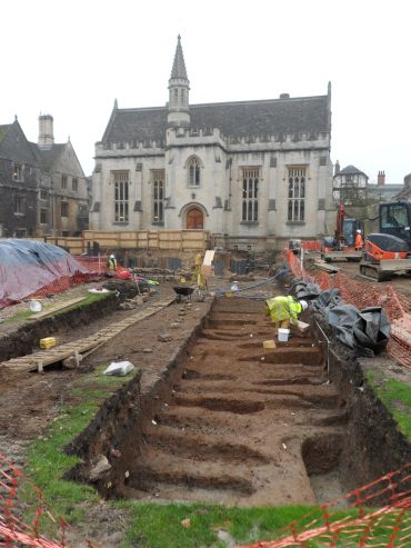 An archaeological site in a courtyard surrounded by buildings. A man in a fluorescent jacket examines a row of oval shaped pits. A building shaped like a chapel is in the background, this is Magdalen College library.