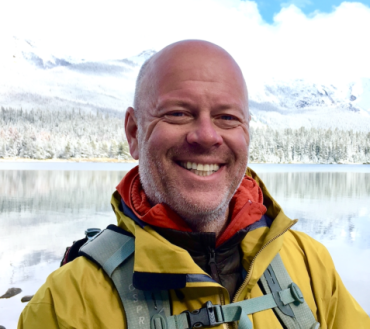 A portrait photograph of Professor Mike Kendall, a middle-aged man with a bald head. He wears an outdoor coat and stands in front of a lake with snow covered mountains in the background.