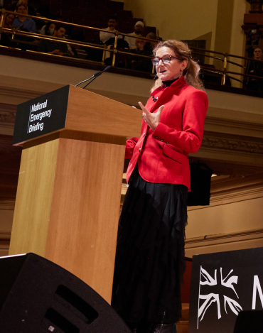 A white woman with blonde hair wearing a red jacket and black skirt stands at a lectern branded 'National Emergency Briefing' 