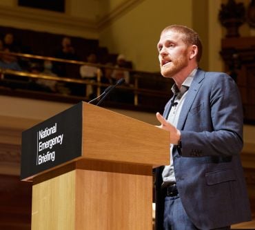 A white man wearing a grey-blue suit speaks at a lectern branded with 'National Emergency Briefing.'