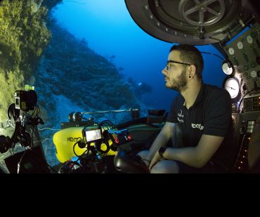 A researcher sits in the driving seat of a submersible, looking out through the windows at the surrounding coral reefs.