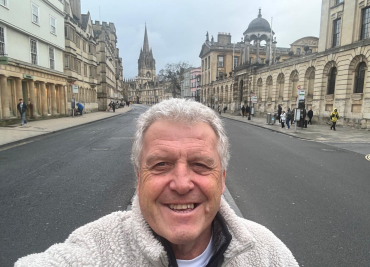 Peter Grindrod, a white man with white hair, standing in Oxford High Street with The Queen’s College and the University Church of St Marys visible.