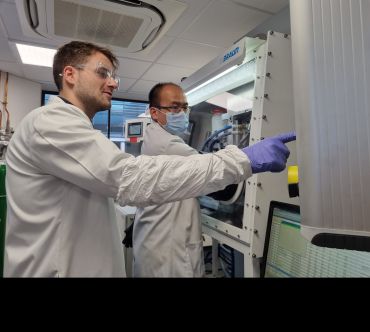 Two male researchers wearing lab coats and latex gloves point to a machine within a laboratory in the University of Oxford’s Department of Materials.