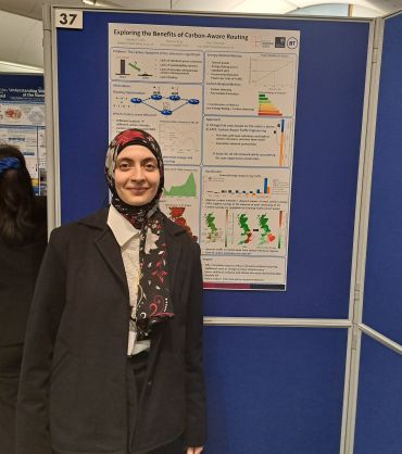 A young lady of Asian descent wearing a headscarf stands in front of a a large poster which summarises her scientific research.