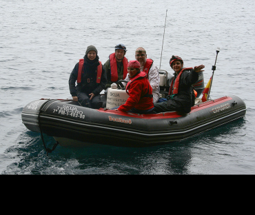 A group of five men and women in an inflatable dinghy on the sea. They are dressed in hardy overalls and smile at the camera.