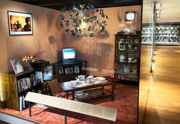 Image depicts a Caribbean living room from the 50s and 60s complete with tea laid out on the coffee table, china cabinet and television on a cabinet