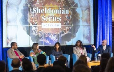 Moderator Dr Julius Grower addresses the panellists on stage with the Sheldonian Series graphic on screen behind them