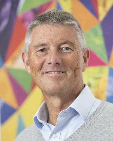 A portrait photograph of Stewart Cole, a middle-aged man with short grey hair. He stands in front of an abstract background made of a repeating shape in different colours. 
