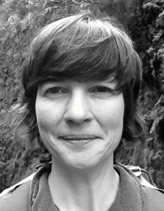 Black and white portrait photo of a woman with shoulder length hair. She stands outside in front of a hedge.