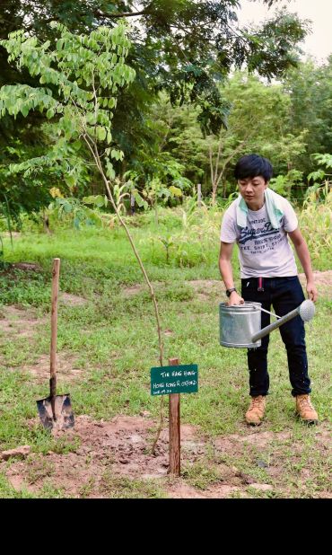 A man holding a watering can stands next to a young tree.