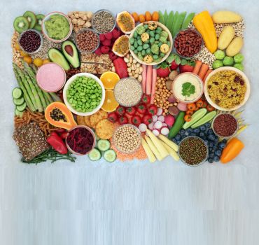 A colourful array of plant-based foods including vegetables, fruit, cereals, grains, nuts, seeds, legumes & dips.