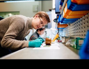 A white man wearing a brown jumper and latex gloves works on electronic equipment on a workbench. 