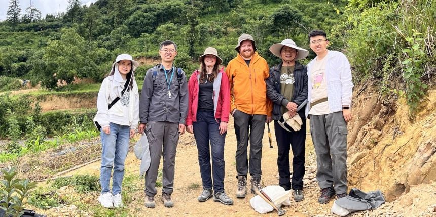 A group of six men and women stand in a rocky environment with a forest in the background.