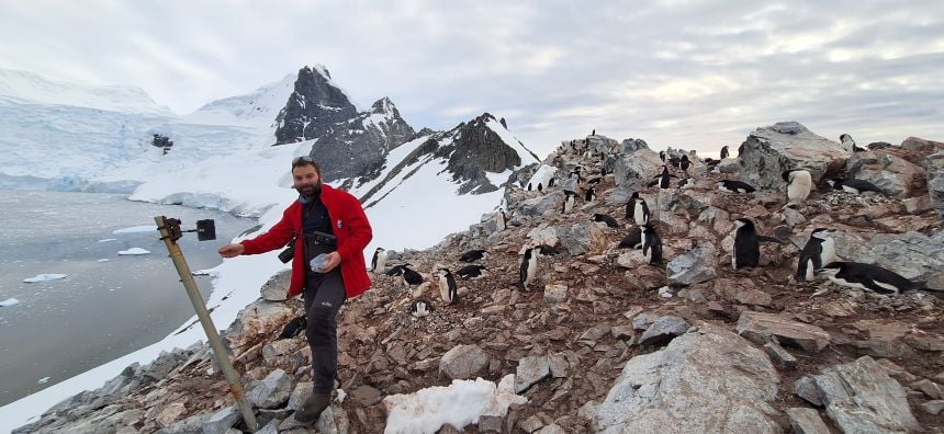 A man wearing a warm red coat stands next to a camera mounted on a tripod on a rocky shoreline, with icy mountains and the Antarctic sea in the background. Around him are several black and white penguins.