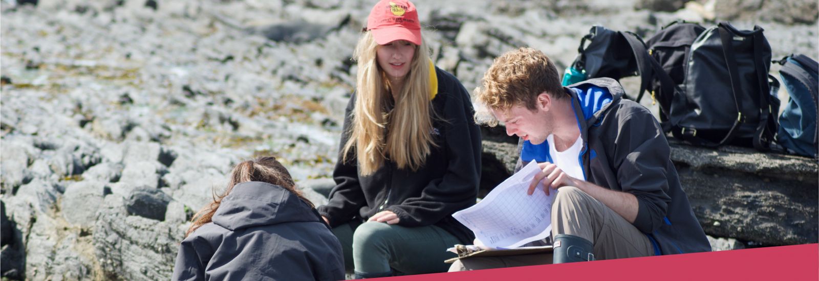 Three students in outdoor clothes record data on a clipboard in a rocky area of Orielton, Wales