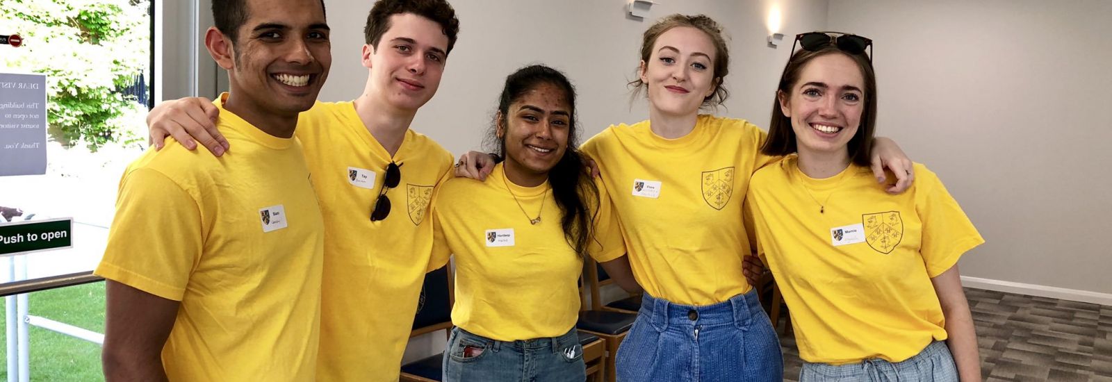A group of five male and female students stand with their arms around each other in the Trinity College lawns pavilion during an open day. They are wearing matching bright yellow t-shirts.