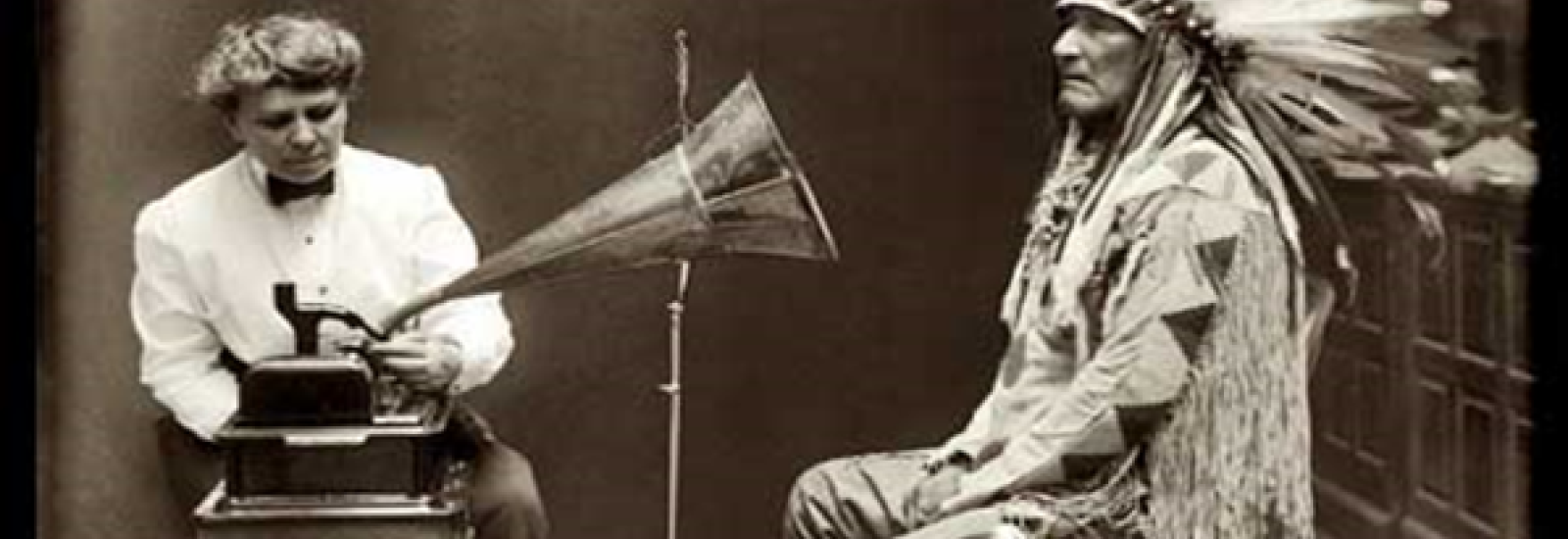 A black and white, historical photograph of a native american, speaking into an antiquitated recording device.