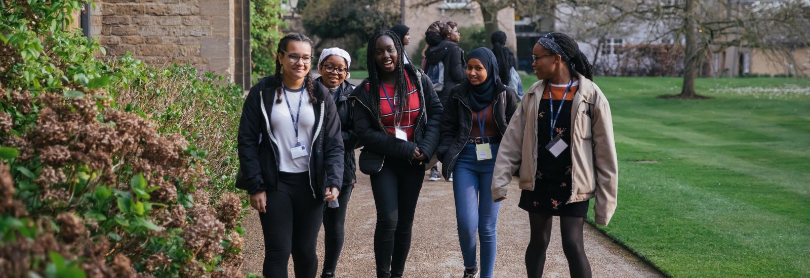 A group of female students wearing lanyards chat and smile while walking on a path in the Trinity College front quad during a school outreach visit.