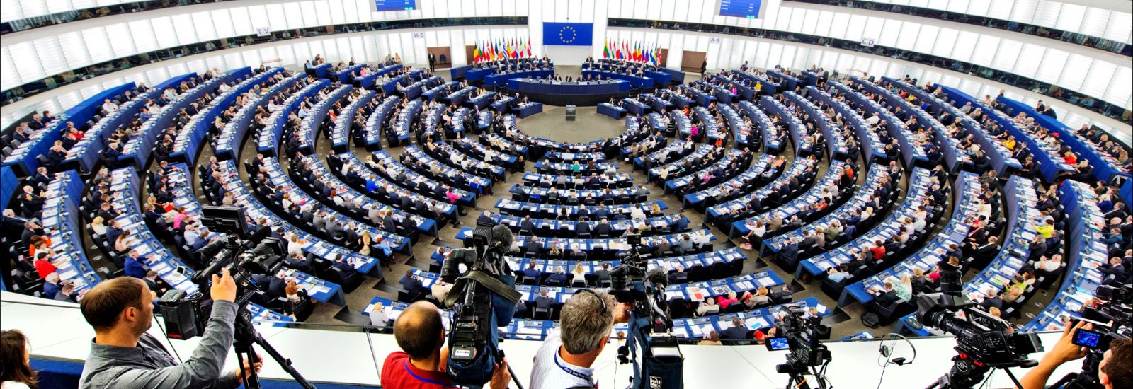 A panoramic view of the interior of the European Parliament, full of parliamentarians ahead of a vote. In the foreground a group of cameramen and photographers position their cameras onto the room. 