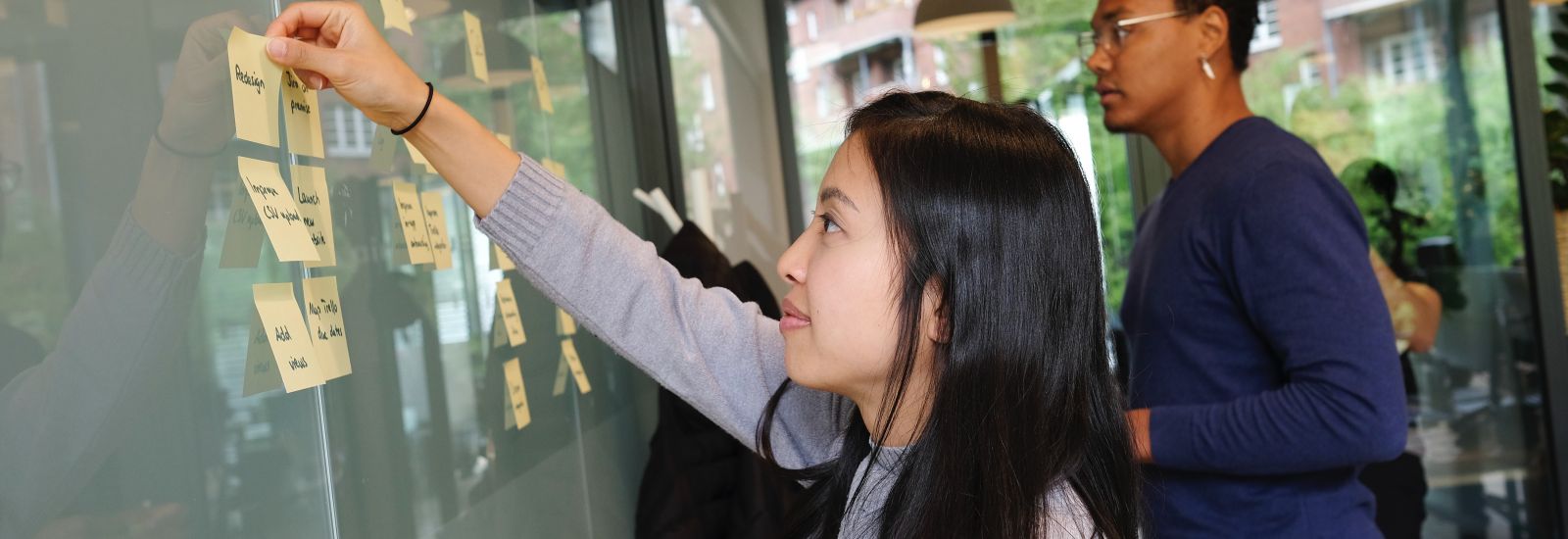 Two young researchers, representing different genders and ethnicities, map out a plan using post-it notes on a whiteboard