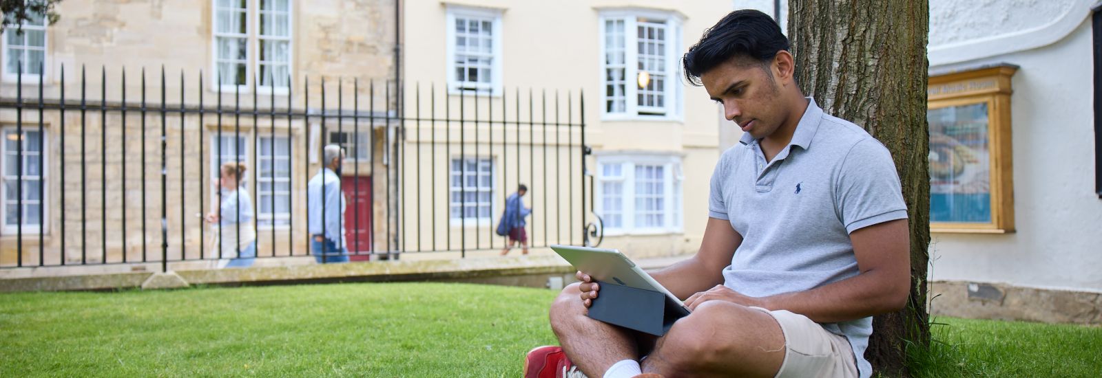 Oxford student with a laptop sat by a tree. 