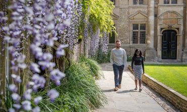 Two students walking in the grounds of St Edmund Hall College next to flowering wisteria. By John Cairns. 