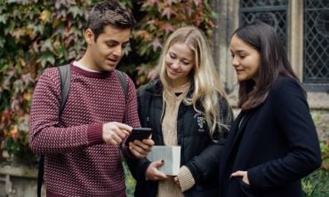 Three students looking at a phone in the grounds of a college